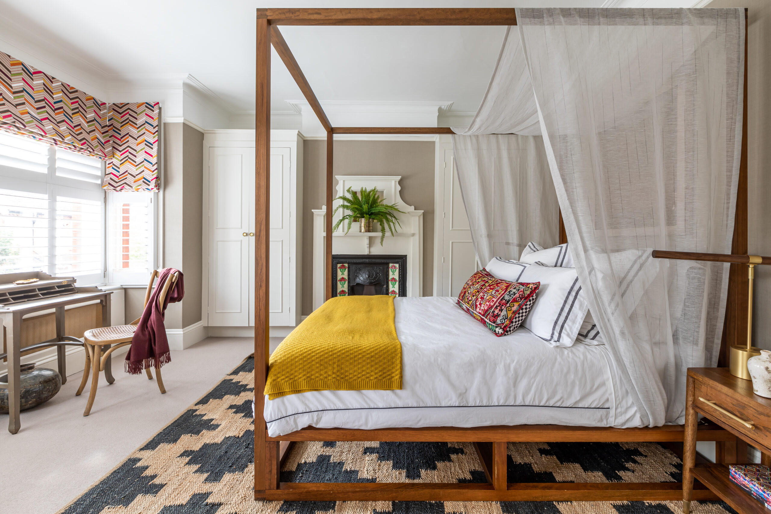 Teak four poster bed with yellow blanket and black and sisal rug. Square bay window with mulitcoloured stitched roman blinds.